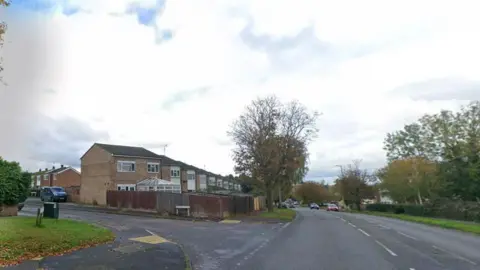 A junction. There are houses on the left side with trees and cars driving across the road 