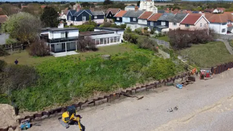 John Fairhall/BBC A modern flat-roofed white house on a grassy cliff in Thorpeness, which runs down to a pebbly beach. Where the cliff meets the beach is a metal fence made up of a row of rust-coloured interlocking sheets of metal. There is a yellow digger on the beach at the bottom left.