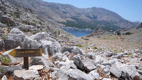 A direction sign on a rocky path in the hills of Pedi (Pedi centre pictured in the distance, right), a small fishing village in Symi, Greece, pointing toward Agia Marina on 8 June