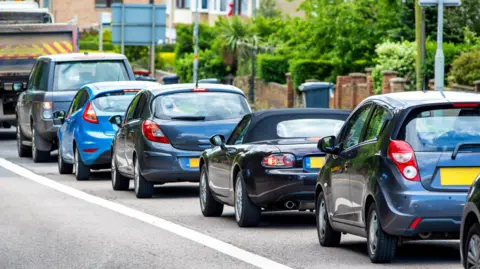 A queue of cars, all of different shapes and sized, queued up on a residential street. 