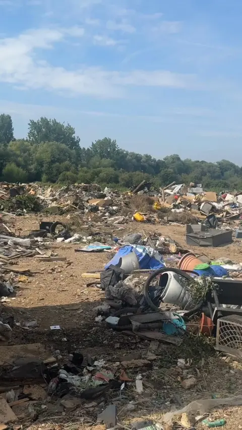 A huge pile of rubbish is seen spread over an area of waste land with a line of trees in the background