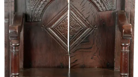 A close-up image of a dark wooden chair. The chair has a noticeable gap in the middle of its back and is carved with elaborate illustrations.