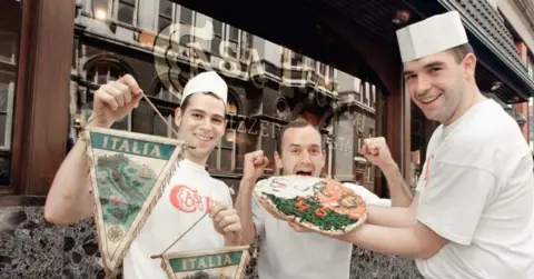 Casa Italia Three chefs wearing white T-shirts with the Casa Italia branding, are holding up a pizza on a plate and some Casa Italia signs. 