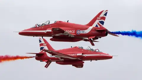 Two Red Arrows jets fly past each other during a display. One is firing out red smoke and the other is letting out blue smoke.