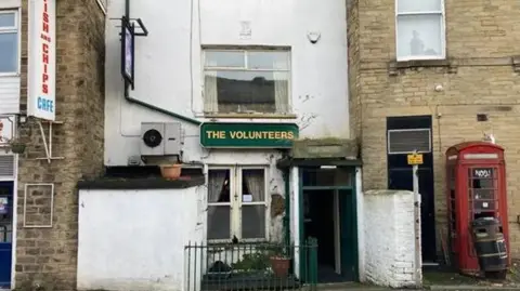 Charles Heslett/BBC The front of a traditional pub called The Volunteers. Picture shows quite a run down building, with shabby windows and door. Also in view is an old rec phone box with the glass in its door broken.