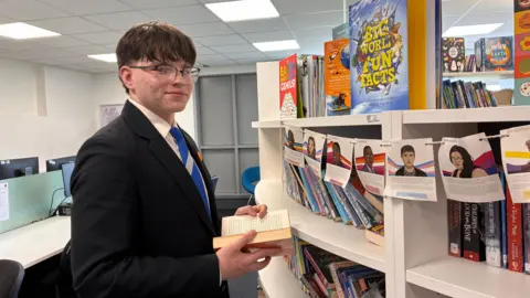 A white teenage boy with brown hair and wearing glasses and school uniform is holding a book in a school library on the left of the picture. He is standing in front of a curved bookcase with computer work stations behind him.
