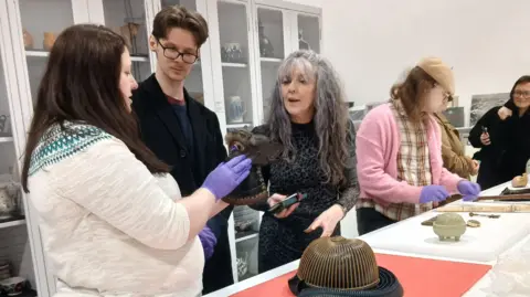 Penlee House Gallery and Museum A group of young people hold objects from the museum's store rooms. A red mat sits on a table with object on it. A woman wearing gloves inspects a piece of art while talking to a young person and a curator.