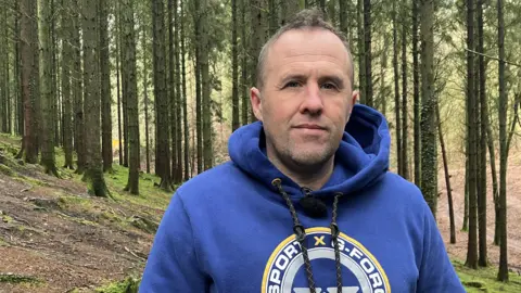 A man is looking at the camera. He is wearing a blue hoodie and has short brow hair. He is stood in a forest.