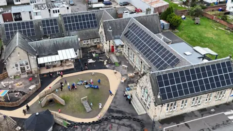 An aerial image of a school with multiple buildings and a central play area. There are children in the playground and several of the roofs are covered with solar panels.