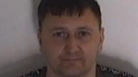 A police mugshot shows a man with short brown hair looking at the camera. He is wearing a dark coloured top.