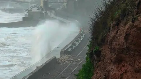 Richard Heiron Waves crash over a sea wall and railway line. Part of the wall is damaged due to the waves. Debris is on the railway track.