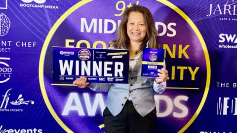 GB Hospitality Marketing A woman with shoulder length light brown hair standing in front of a purple, gold and white backdrop that has writing on it. It reads "Midlands Food and Drink Hospitality Awards." The woman is holding a glass award that says "winner" and a board that reads "winner"