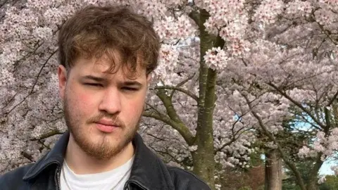 Family Handout William Hewes who has brown hair and a beard and is wearing a leather jacket in front of some light pink cherry blossom trees