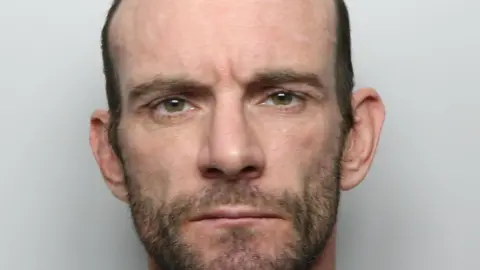 A police custody picture of a man with balding hair and a short beard.