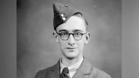 Young man wearing glasses and a military uniform. He is looking into the camera with a serious expression and wearing his RAF hat tilted to the side of his head.