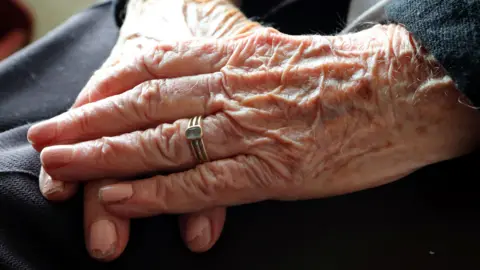 File image showing an elderly woman, wearing chipped pale pink nail polish and a gold ring with a blue stone, crossing her hands on her lap