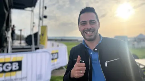 Telmo smiles at the camera as he gives a thumbs up. He has a black jacket on with a blue polo t-shirt. Behind him is the music stage and the sun is shining down on the field.