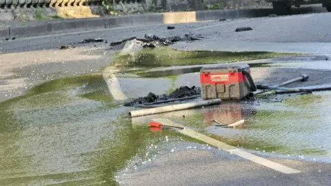 John Parish A close-up of the water pooled on the road surface. There appears to be damage to the road surface.