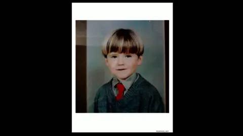 Family photo Oran Doherty's school photo - he has short, blonde hair styled in a bowl cut.  He is wearing a grey school shirt and jumper and a red tie.