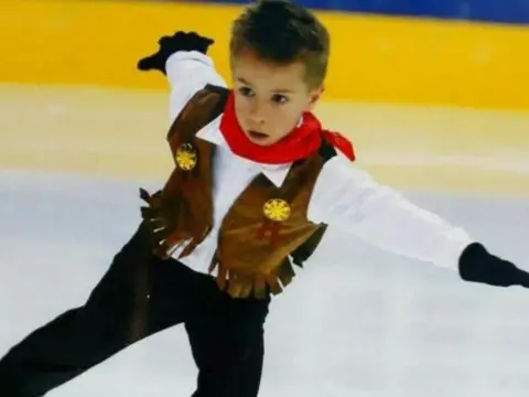 Karen Brennan A young Edward stretches out in a star shape while on the ice. He is wearing a cowboy costume with a red neckerchief. There is a look of deep concentration on his dace.