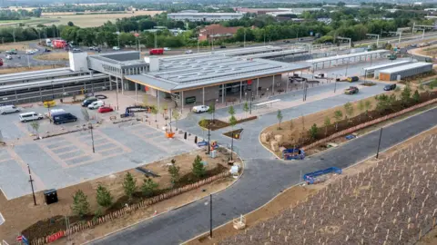 Beaulieu Park station has a large building next to three platforms, which have tall grey covers over them to protect passengers from the weather. There is a bus interchange and a car park. The station is surrounded by roads and lots of trees.