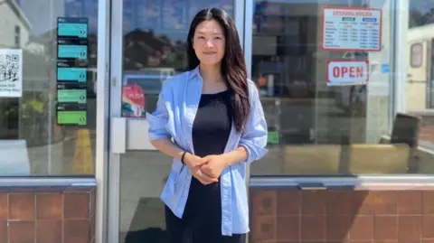BBC/Angela Ferguson A woman with long black hair and wearing a black dress and light blue blouse is smiling as she stands in front of the entrance to the Rainbow 2000 restaurant.
