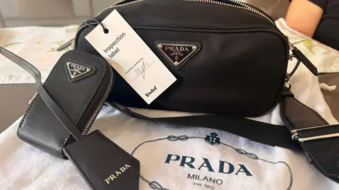 A fake black nylon Prada handbag with a phone case and a white cotton bag with the words 'Prada Milano Dal 1913' on it. It has a white ticket tied to the strap which says 'inspection label' and 'expertly verified by' with a signature and the Vinted logo.