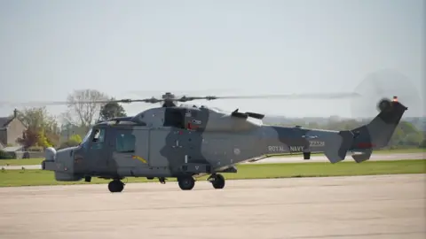 A camouflaged Wildcat military helicopter with rotors whirring on a the tarmac runway taking off at RNAS Yeovilton 