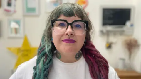 A close-up shot of Kat looking into the camera wearing large black-rimmed glasses, with the background out of focus.