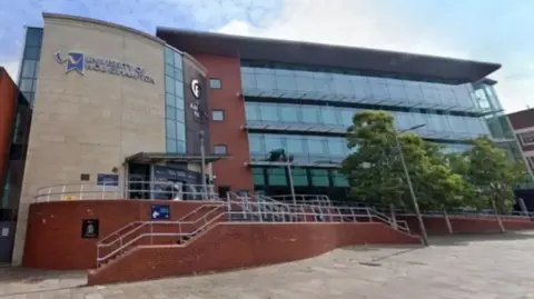 The front of the university is glass, red brick and stone. There is a railed walkway up to the entrance and some trees on the right hand side.