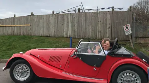 A woman in a white T-shirt is in the passenger seat of an open-top vintage red car. She is laughing and smiling while a man next to her drives.