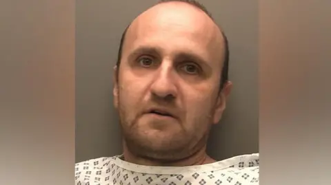 Custody photograph of Veselin Dudenski. He has short black hair and beard and is wearing a white top with black squares on.