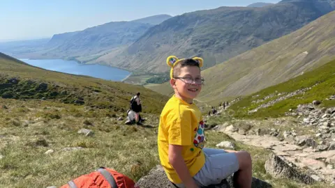 A boy with brown hair and glasses in a yellow T-shirt and white shorts sat on a rock, part way up a large hill which can be seen stretching down to a lake below and there are other large hills in the distance
