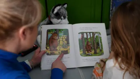 A shot from over the shoulder of a woman pointing to a book and a child reading the book. The woman has blonde hair tied back and is wearing a blue long sleeve top. The child has long brown hair and is wearing a white t-shirt. The book is The Gruffalo and has illustrations and words on both pages. 