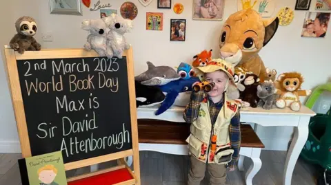 Samantha Evans-Browning A little boy dressed in safari gear as he emulates his hero Sir David Attenborough. Next to him is a board that says "2nd March 2023. World Book Day. Max is Sir David Attenborough." Behind him a soft cuddly toys of various sizes and animals, including a sloth, koala bears, a lion, dolphin, orca, rhino and a monkey.