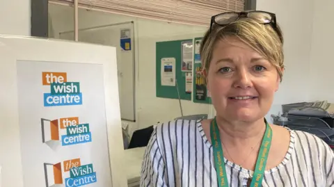 Rebekah Wilson wearing a striped top standing next to a wish centre sign in their office building