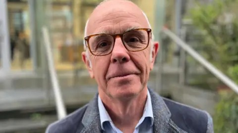 BBC Dr Steve Evans - A bald man with white hair on the side of his heard, wearing brown glasses. he has a grey suit on with a blue shirt. 