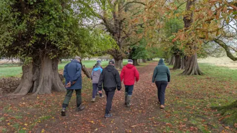 National Trust Images/Paul Harris A group of walkers in coats, hats and wellies wandering along a lane surrounded by trees. There are quite a few orange leaves, suggesting it is autumn.