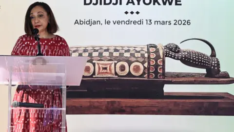 Ivorian Minister of Culture and Francophonie Françoise Remarck talks at a podium in front of a picture of the ornately carved wooden drum