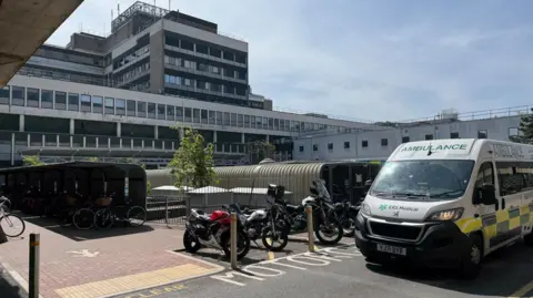 BBC/Steve Hubbard The exterior of Addenbrooke's Hospital, a white and grey building, with a parked ambulance and motorcycles in the foreground