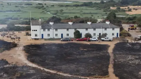 Reuters A row of about six white terraced cottages. In front of them is blackened earth. Behind is green fields and trees untouched by fire. The photo is taken from above.