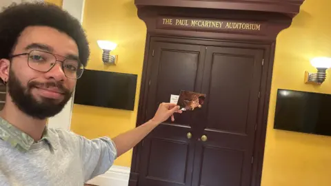 A male student at LIPA in Liverpool holding the postcard in front of a door which reads 'the paul mccartney auditorium'