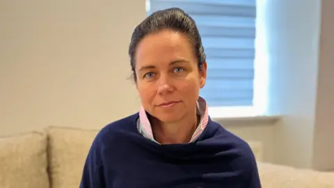 Digby Brown A woman in a blue jumper with a pink shirt underneath looks into the camera while seated on a sofa