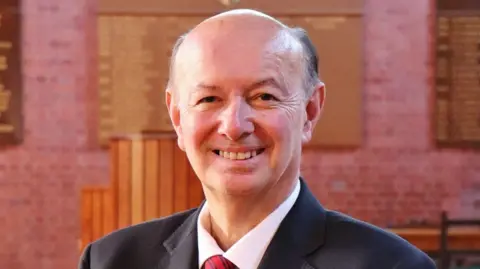 Ellesmere College A man with thinning grey hair and a dark blue suite with a red tie in a room with brick walls