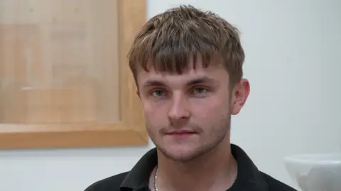 Jamie Niblock/BBC A young man smiles slightly at the camera. He has light brown hair which covers his forehead as well as some dark stubble. He is wearing a black polo top and a silver chain around his neck.