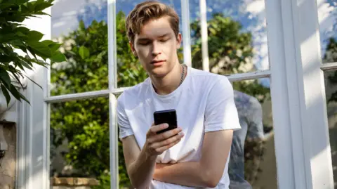 Getty Images Młody mężczyzna stoi przed domem i patrzy w telefon, a w oknie za nim odbija się drzewo