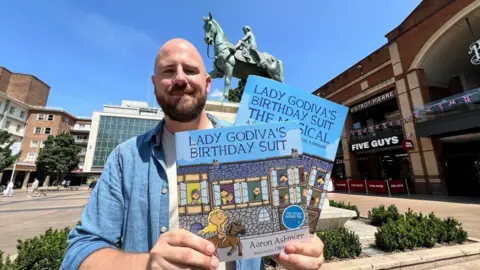 A bald man in a blue shirt and white top. He is holding two books which are called "Lady Godiva's Birthday Suit" and standing in the middle of a city centre. The sky is bright blue in the background and there are shops and restaurants behind him.