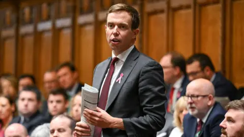 Reuters/House of Commons A man, wearing a grey suit stands among a group of people, mainly men, in the House of Commons. He is holding a piece of paper on his hands.