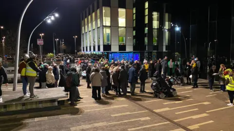 A crowd in coats and hats congregate outside the Corby Cube