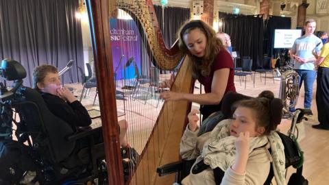 SEND pupils enjoying listening to the harp, with woman wearing red top playing it in big concert hall. 